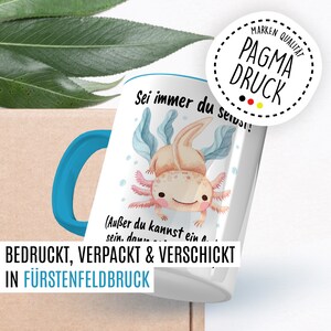 May include: A white ceramic mug with a blue handle features a watercolor illustration of an axolotl with the text "Sei immer du selbst!" (Au&szlig;er du kannst ein Axolotl sein. dann..."  The mug is printed, packaged, and shipped from F&uuml;rstenfeldbruck.