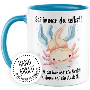 May include: White ceramic mug with a blue rim and handle. The mug features a watercolor illustration of an axolotl with the text "Sei immer du selbst!" and "er du kannst ein Axolotl, dann sei ein Axolotl". A white circle sticker with black text reads "HAND ARBEIT IN DEUTSCHLAND BEDRUCKT".
