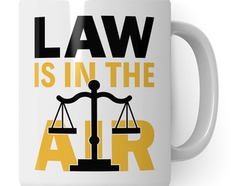 Może przedstawiać: Biały kubek ceramiczny z czarno-złotym wzorem. Kubek zawiera napis "LAW IS IN THE AIR" z czarnym symbolem wagi sprawiedliwości.