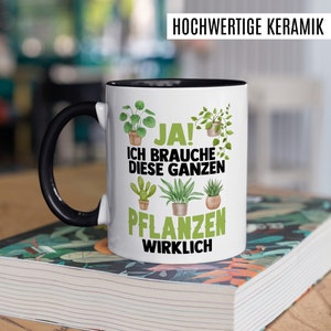 May include: White ceramic mug with a black handle and rim. The mug has a green and black design with the text "JA! ICH BRAUCHE DIESE GANZEN PFLANZEN WIRKLICH" on it.