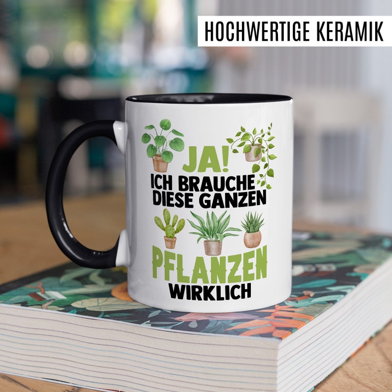 May include: White ceramic mug with a black handle and rim. The mug has a green and black design with the text "JA! ICH BRAUCHE DIESE GANZEN PFLANZEN WIRKLICH" on it.