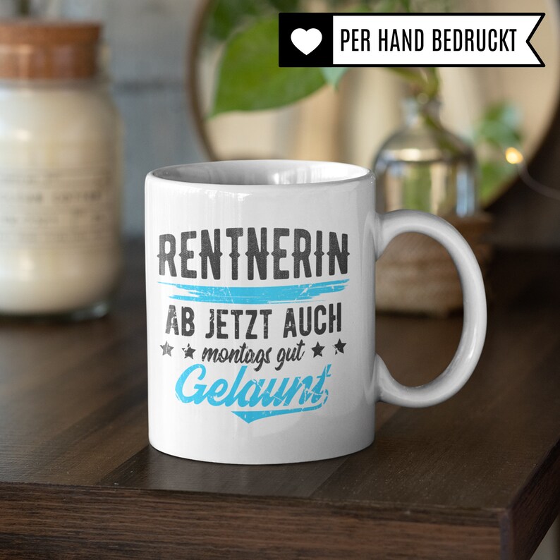 May include: White ceramic mug with a black and blue design. The mug says "Rentnerin ab jetzt auch montags gut gelaunt". The mug is on a wooden table.