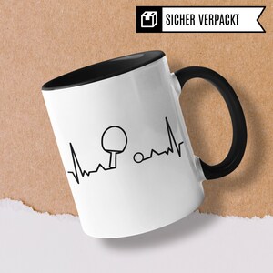 May include: White ceramic mug with a black handle and a black interior. The mug features a black and white graphic of a table tennis paddle and ball with a heartbeat line. The text "SICHER VERPACKT" is on the top of the mug.