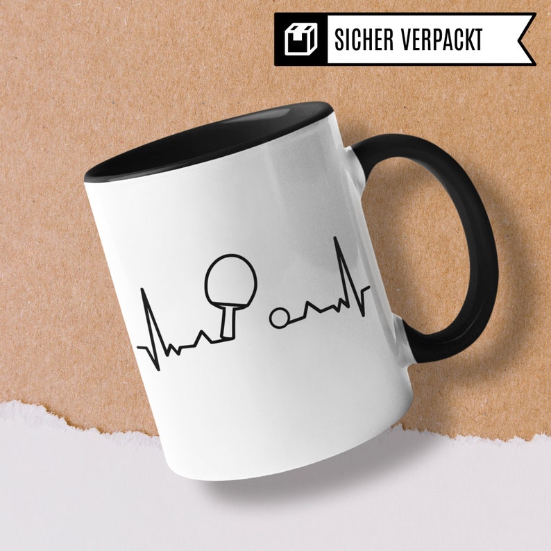 May include: White ceramic mug with a black handle and a black interior. The mug features a black and white graphic of a table tennis paddle and ball with a heartbeat line. The text "SICHER VERPACKT" is on the top of the mug.