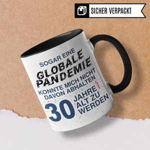 May include: A white coffee mug with a black handle and black rim. The mug has a colorful confetti design and black text that reads "SOGAR EINE GLOBALE PANDEMIE KONNTE MICH NICHT DAVON ABHALTEN 30 JAHRE ALT ZU WERDEN".