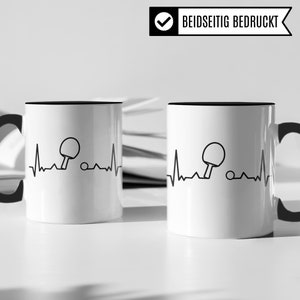 May include: Two white ceramic mugs with black rims. Each mug features a black and white illustration of a heartbeat with a table tennis bat and ball inside the heartbeat. The text "BEIDSETIG BEDRUCKT" is visible on a white tag.