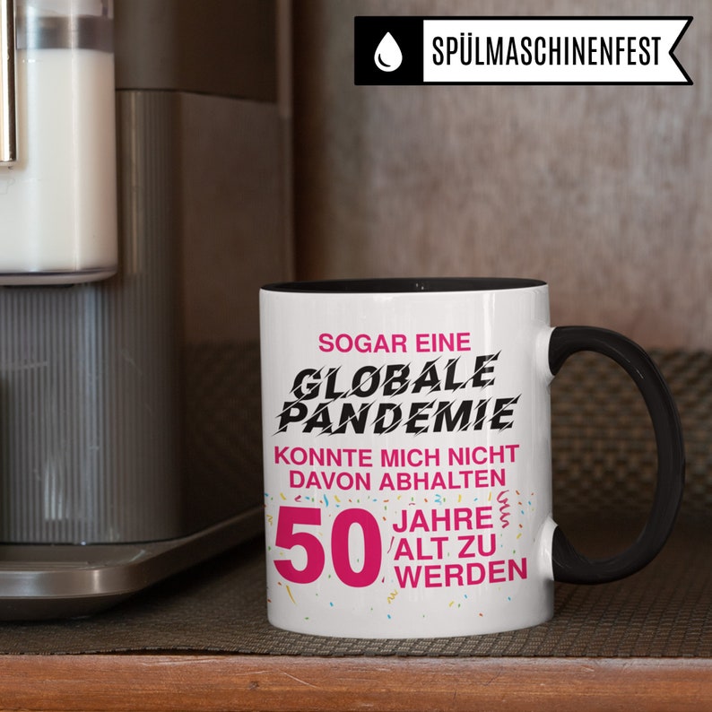 May include: A white coffee mug with a black handle and black rim. The mug has a pink and black graphic that says "Sogar eine Globale Pandemie Konnte mich nicht davon abhalten 50 Jahre alt zu werden".