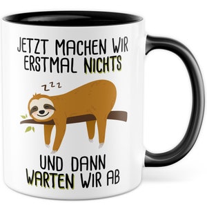 Könnte beinhalten: Weiße Keramiktasse mit schwarzem Rand und Henkel. Die Tasse zeigt ein Cartoon-Faultier, das an einem Ast hängt, mit dem Text "Jetzt machen wir erstmal nichts und dann warten wir ab" und einem schlafenden ZZZ-Symbol über dem Faultier.