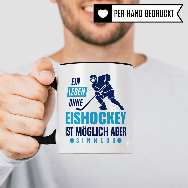 Pu&ograve; includere: Tazza in ceramica bianca con bordo e manico neri. La tazza presenta una grafica blu e bianca di un giocatore di hockey con il testo "Ein Leben Ohne Eishockey Ist M&ouml;glich Aber Sinnlos".