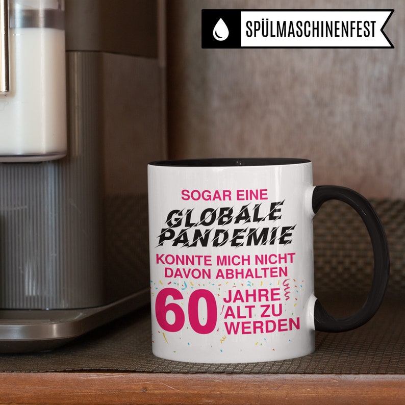 May include: White coffee mug with a black handle. The mug has black text that reads "SOGAR EINE GLOBALE PANDEMIE KONNTE MICH NICHT DAVON ABHALTEN 60 JAHRE ALT ZU WERDEN". The text is surrounded by pink confetti.