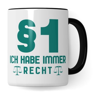 Könnte beinhalten: Weiße Keramiktasse mit schwarzem Rand und Henkel. Die Tasse hat ein grünes Paragraphenzeichen und die Zahl 1 darauf gedruckt. Der Text "Ich habe immer Recht" ist unter der Zahl gedruckt, mit einer Waage auf jeder Seite des Wortes "Recht".