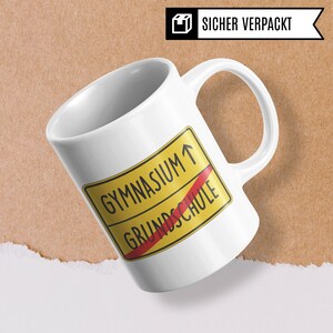 Może przedstawiać: Biały kubek ceramiczny z ż&oacute;łto-czerwonym znakiem drogowym, na kt&oacute;rym widnieje napis "Gymnasium" ze strzałką skierowaną w g&oacute;rę i "Grundschule" z przekreśloną czerwoną linią.
