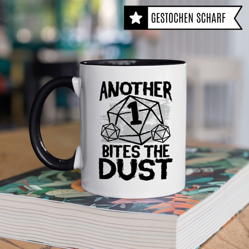 May include: White ceramic mug with a black handle and black rim. The mug has a graphic of a dice with the number one on it and the text "Another 1 Bites The Dust".