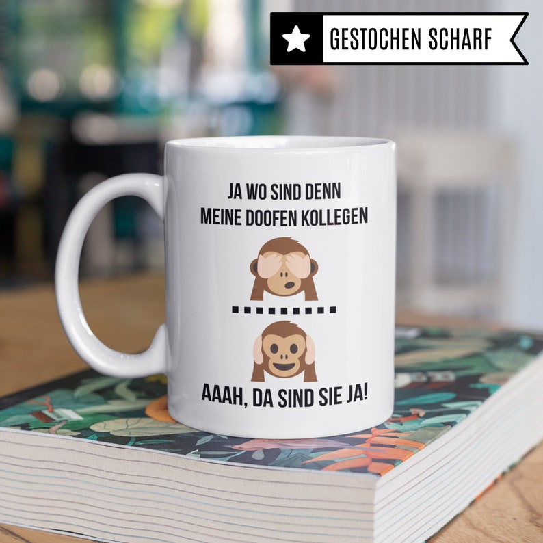 May include: White ceramic coffee mug with a black and brown monkey graphic and the text "JA WO SIND DENN MEINE DOOFEN KOLLEGEN" and "AAAH, DA SIND SIE JA!" printed on it.