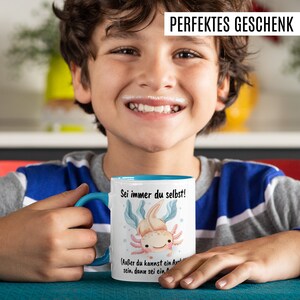 May include: A white ceramic mug with a blue rim and handle. The mug features a cartoon axolotl with the text "Sei immer du selbst!" and "(Au&szlig;er du kannst ein Axolotl sein, dann sei ein Axolotl)".
