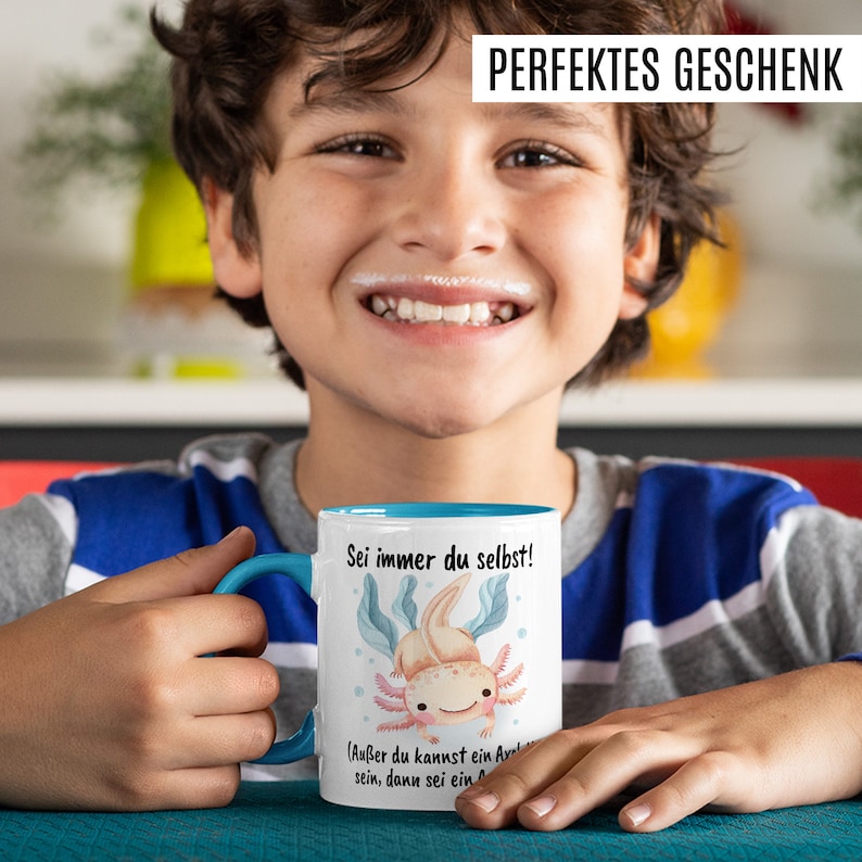 May include: A white ceramic mug with a blue rim and handle. The mug features a cartoon axolotl with the text "Sei immer du selbst!" and "(Au&szlig;er du kannst ein Axolotl sein, dann sei ein Axolotl)".