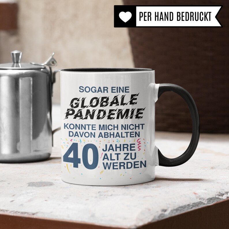 May include: White ceramic mug with a black rim and handle. The mug has black text that reads "Sogar eine Globale Pandemie Konnte mich nicht davon abhalten 40 Jahre alt zu werden".