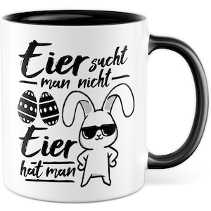 Op de afbeelding: Witte keramische mok met een zwarte rand en handvat. De mok heeft een zwart-witte illustratie van een konijn met een zonnebril met de tekst "Eier sucht man nicht - Tier hat man" erop gedrukt.