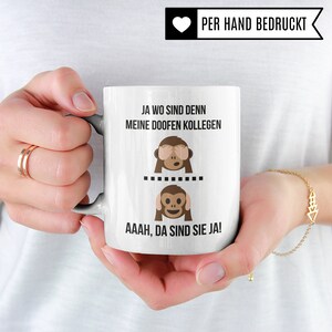 May include: White ceramic mug with a black and white illustration of two monkeys. The top monkey is covering its eyes and the bottom monkey is covering its ears. The text on the mug reads "Ja wo sind denn meine doofen Kollegen" and "Aahh, da sind sie ja!"