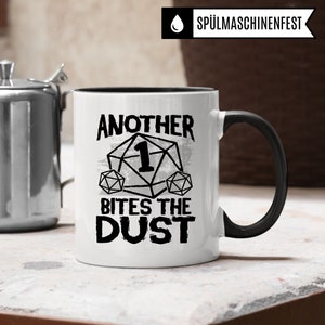 May include: White ceramic mug with a black rim and handle. The mug has a black and white graphic of a dice with the number one on it and the text "Another Bites The Dust".