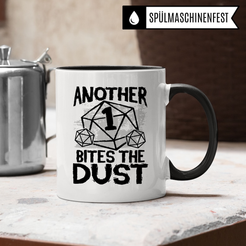 May include: White ceramic mug with a black rim and handle. The mug has a black and white graphic of a dice with the number one on it and the text "Another Bites The Dust".