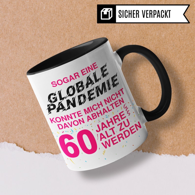May include: A black and white mug with a black handle. The mug has a white background with black text that reads "SOGAR EINE GLOBALE PANDEMIE KONNTE MICH NICHT DAVON ABHALTEN 60 JAHRE ALT ZU WERDEN". The text "60" is in pink. There are colorful confetti sprinkles on the mug.