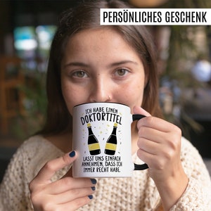 May include: White coffee mug with a black rim and handle. The mug has a black and gold design with the text "Ich habe einen Doktortitel. Lasst uns einfach annehmen, dass ich immer recht habe."