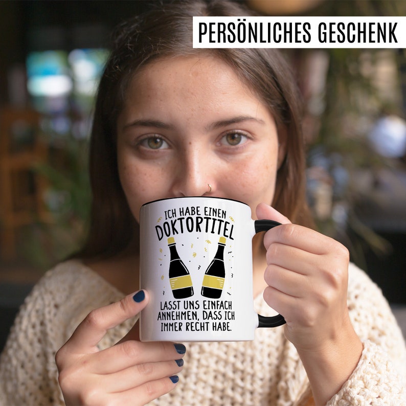 May include: White coffee mug with a black rim and handle. The mug has a black and gold design with the text "Ich habe einen Doktortitel. Lasst uns einfach annehmen, dass ich immer recht habe."