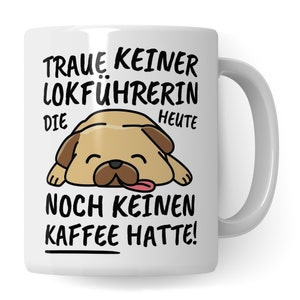 Pode incluir: Caneca de cerâmica branca com uma ilustração de um cão pug de desenho animado. A caneca tem texto preto que diz "Traue keiner Lokführerin die heute noch keinen Kaffee hatte!"