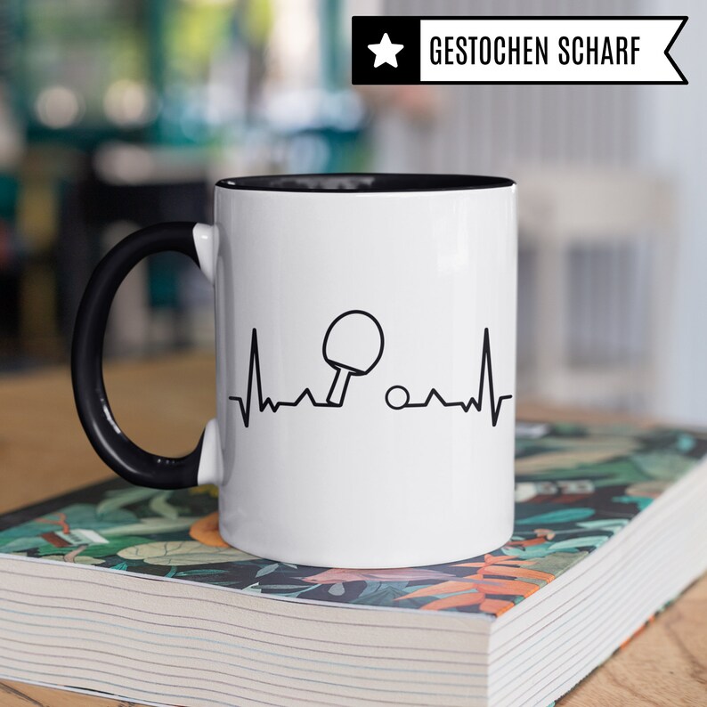May include: White ceramic mug with a black handle and a black rim. The mug features a black and white graphic of a table tennis bat and ball with a heartbeat line. The text "GESTOCHEN SCHARF" is on a black star in the top right corner of the image.