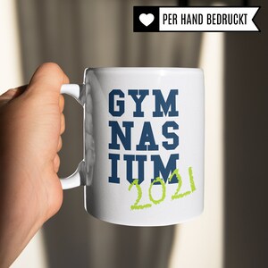 Pu&ograve; includere: Tazza in ceramica bianca con un design blu e verde. La tazza dice "GYMNASIUM 2021".