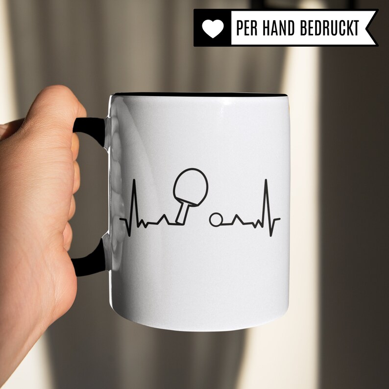 May include: White ceramic mug with a black handle and a black rim. The mug features a black and white graphic of a table tennis bat and ball with a heartbeat line. The text "PER HAND BEDRUCKT" is printed on a white heart.