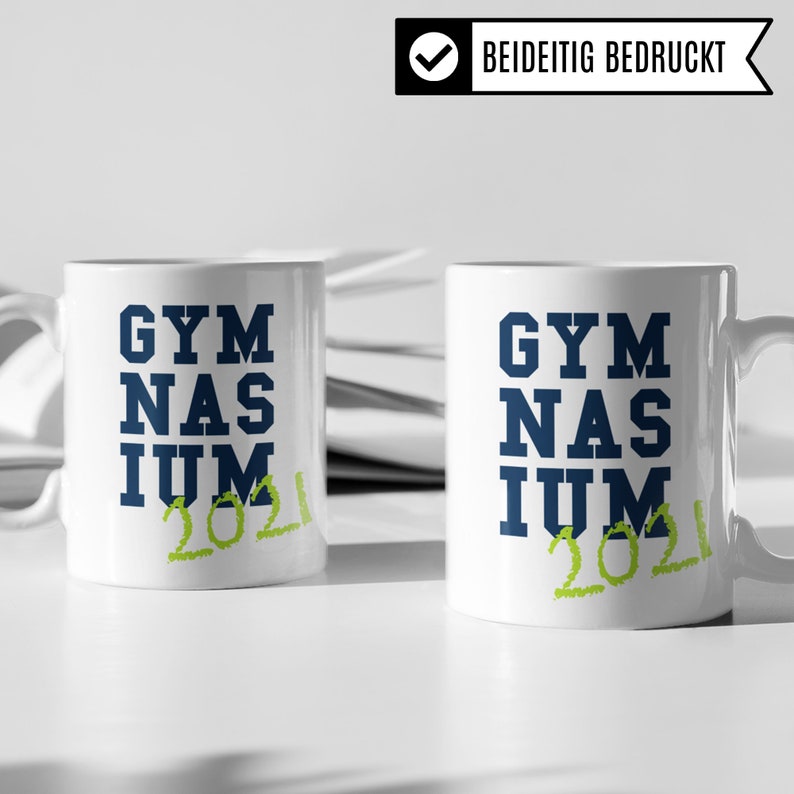 Pu&ograve; includere: Due tazze in ceramica bianca con testo blu che recita "GYMNASIUM 2021" in verde. Le tazze sono su una superficie bianca.