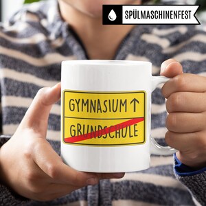 Może przedstawiać: Biały kubek ceramiczny z ż&oacute;łto-czerwonym znakiem, na kt&oacute;rym widnieje napis "Gymnasium &uarr;" z przekreślonym czerwonym paskiem "Grundschule".