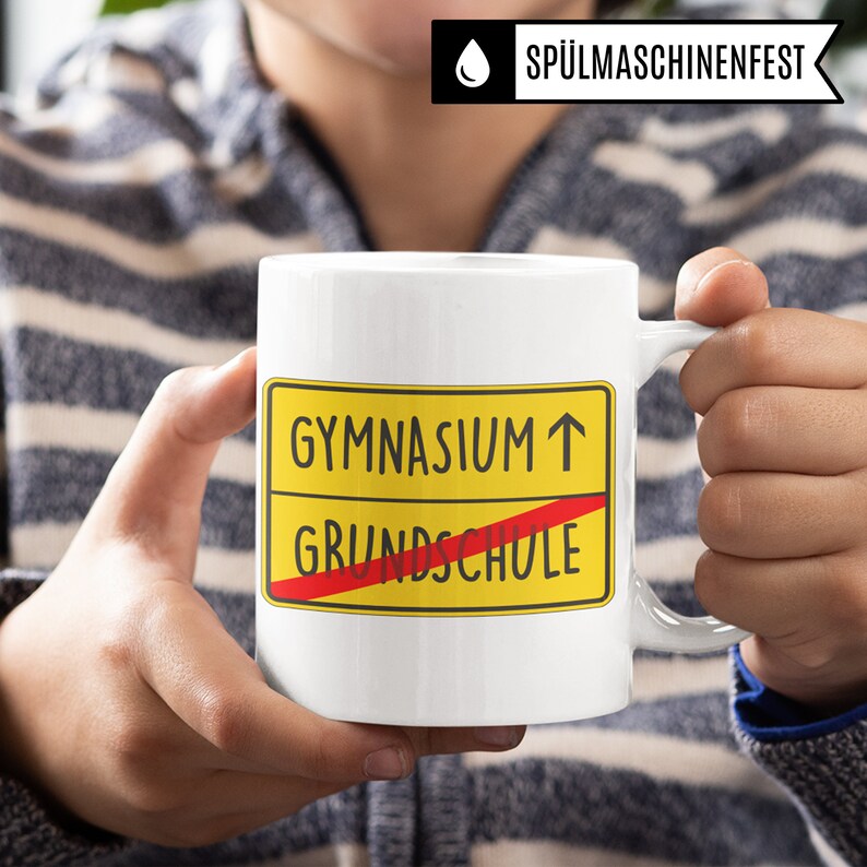 Może przedstawiać: Biały kubek ceramiczny z ż&oacute;łto-czerwonym znakiem, na kt&oacute;rym widnieje napis "Gymnasium &uarr;" z przekreślonym czerwonym paskiem "Grundschule".