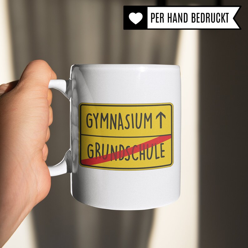 Może przedstawiać: Biały kubek ceramiczny z ż&oacute;łtym i czerwonym znakiem, na kt&oacute;rym widnieje napis "Gymnasium &uarr;" z przekreślonym czerwonym paskiem "Grundschule". Kubek jest ręcznie malowany.