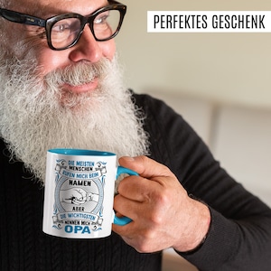 Peut inclure: Une tasse en céramique blanche avec un bord et une poignée bleus. La tasse a un motif circulaire bleu avec du texte blanc qui dit "Die meisten Menschen rufen mich beim Namen aber die wichtigsten nennen mich Opa".