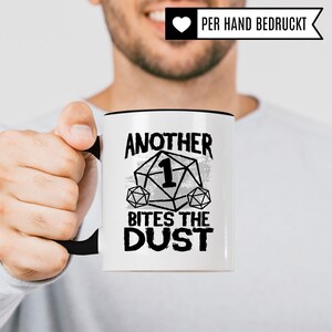 May include: White ceramic mug with a black rim and handle. The mug has a black and white graphic of a dice with the number one on it and the text "Another 1 Bites The Dust".