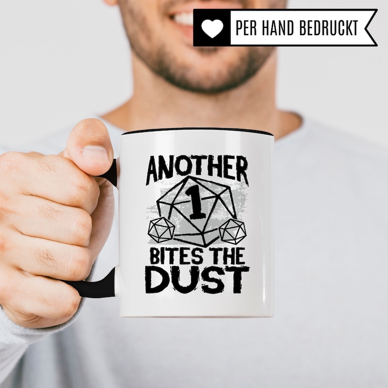 May include: White ceramic mug with a black rim and handle. The mug has a black and white graphic of a dice with the number one on it and the text "Another 1 Bites The Dust".