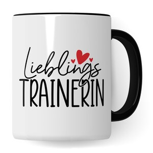 Puede incluir: Taza de cerámica blanca con borde y asa negros. La taza tiene el texto "Lieblings Trainerin" en escritura negra con tres corazones rojos.