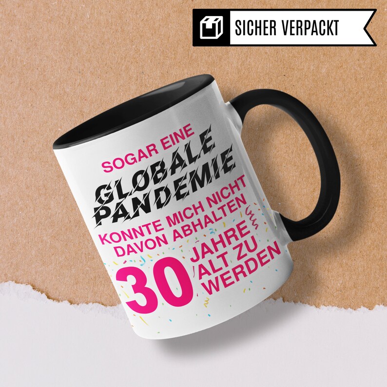 May include: A black and white mug with a black handle. The mug has a white background with black text that reads "Sogar eine Globale Pandemie Konnte mich nicht davon abhalten 30 Jahre alt zu werden". The text "30" is in pink. The mug is sitting on a brown surface.