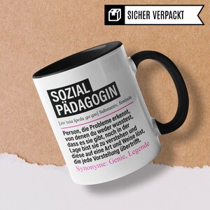 May include: White ceramic mug with a black handle and black interior. The mug has a black and white design with the text "SOZIAL P&Auml;DAGOGIN [zotsia:lpeda go:gin] Substantiv, feminin Person, die Probleme erkennt, von denen du weder wusstest, dass es sie gibt, noch in der Lage bist sie zu verstehen und diese auf eine Art und Weise l&ouml;st, die jede Vorstellung &uuml;bertrifft. Synonyme: Genie, Legende" printed on the front.