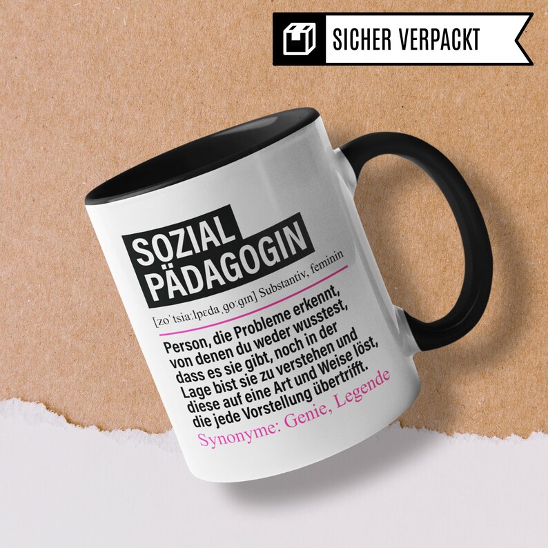May include: White ceramic mug with a black handle and black interior. The mug has a black and white design with the text "SOZIAL P&Auml;DAGOGIN [zotsia:lpeda go:gin] Substantiv, feminin Person, die Probleme erkennt, von denen du weder wusstest, dass es sie gibt, noch in der Lage bist sie zu verstehen und diese auf eine Art und Weise l&ouml;st, die jede Vorstellung &uuml;bertrifft. Synonyme: Genie, Legende" printed on the front.