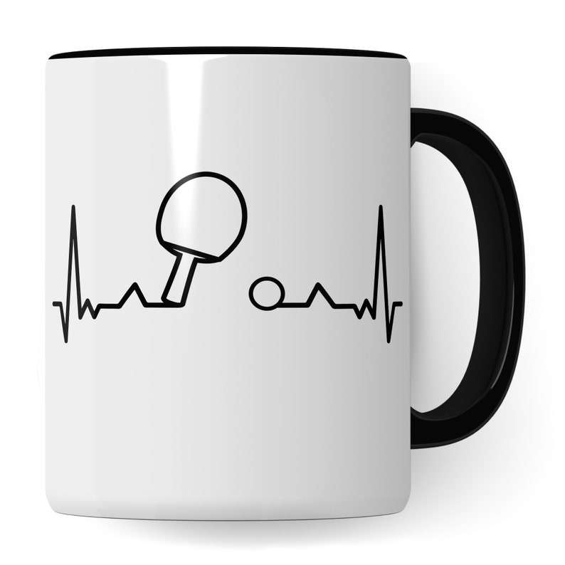 May include: White ceramic coffee mug with a black handle and a black outline. The mug features a black and white illustration of a table tennis bat and ball with a heartbeat line.