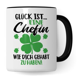 May include: White ceramic mug with a black rim and handle. The mug has a black graphic that says "Glück ist... eine Chefin wie dich gehabt zu haben!" with four green four-leaf clovers.