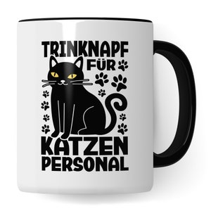 Può includere: Tazza in ceramica bianca con bordo e manico neri. La tazza presenta un gatto nero con occhi gialli e impronte di zampe. Il testo "TRINKNAPF FÜR KATZEN PERSONAL" è stampato sulla tazza.