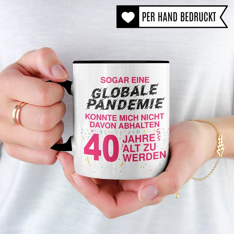 Puede incluir: Taza de cer&aacute;mica blanca con asa negra. La taza tiene un dise&ntilde;o rosa y negro con el texto "SOGAR EINE GLOBALE PANDEMIE KONNTE MICH NICHT DAVON ABHALTEN 40 JAHRE ALT ZU WERDEN".