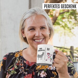 Peut inclure: Une tasse en c&eacute;ramique blanche avec un motif noir et blanc. La tasse a une bordure et une poign&eacute;e roses. Le motif repr&eacute;sente un &acirc;ne sur le c&ocirc;t&eacute; gauche avec le texte "Andere Schwiegerm&uuml;tter" et une licorne sur le c&ocirc;t&eacute; droit avec le texte "Du".