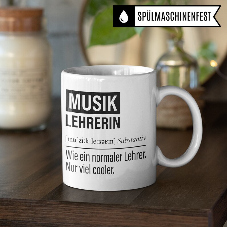 May include: White ceramic mug with a black design that says "MUSIK LEHRERIN [mu'zi:k'le:bərın] Substantiv Wie ein normaler Lehrer. Nur viel cooler."