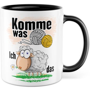 Könnte beinhalten: Weiße Keramiktasse mit schwarzem Rand und Henkel. Die Tasse zeigt ein Cartoon-Schaf mit einer Blume im Maul, zwei Wollknäuel und den Text "Komme was ich das".